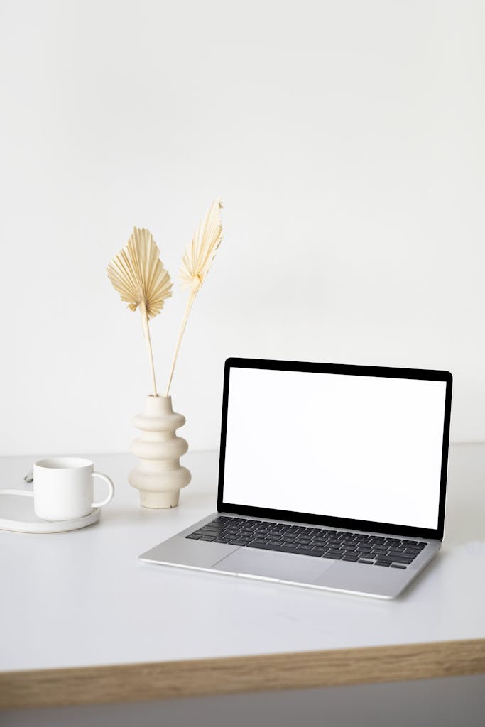 A clean and simple workspace setup with a laptop, ceramic vase, and coffee cup on a wooden table.